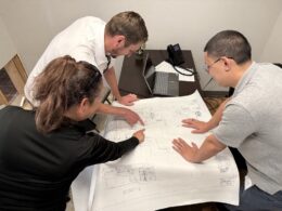 Three people review building layout plans on a desk.